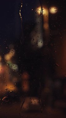 Pencere yüzeyine düşen yağmur damlaları. HD döngü dikey video animasyonu. Gece şehri ve sokak ışıklarıyla manzara. Bulanık camlara akan su damlaları.
