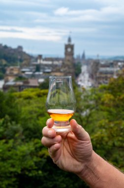 Hand holding glass of single malt scotch whisky and view from Calton hill to park and old parts of Edinburgh city in rainy summer day, Scotland, UK