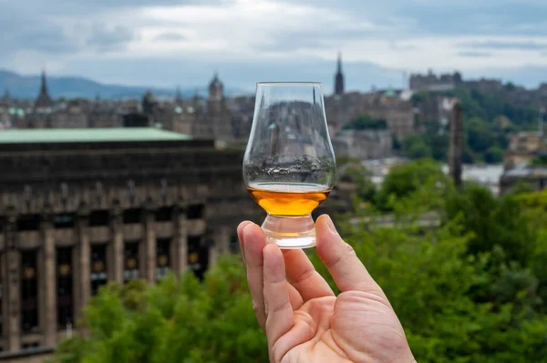 stock image Hand holding glass of single malt scotch whisky and view from Calton hill to park and old parts of Edinburgh city in rainy summer day, Scotland, UK