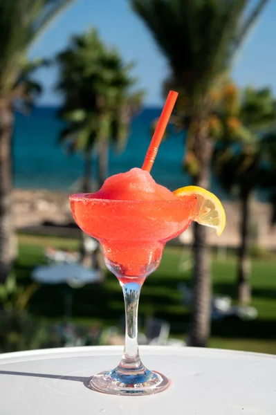 Colourful frozen pink Strawberry daiquiri cocktail drink served in glass at outdoor cafe overlooking blue sea and palm trees, relax and holidays at sea