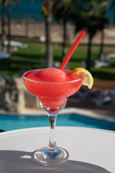 Colourful frozen pink Strawberry daiquiri cocktail drink served in glass at outdoor cafe overlooking blue pool, sea and palm trees, relax and holidays at sea