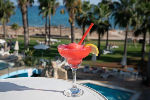 Colourful frozen pink Strawberry daiquiri cocktail drink served in glass at pool bar overlooking blue pool, sea and palm trees, relax and holidays at sea