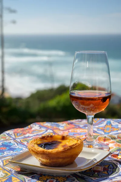Portugal\'s traditional food and drink, glass of porto wine or muscatel de setubal, sweet dessert Pastel de nata egg custard tart pastry served with view on blue Atlantic ocean near Sintra in Lisbon area, Portugal