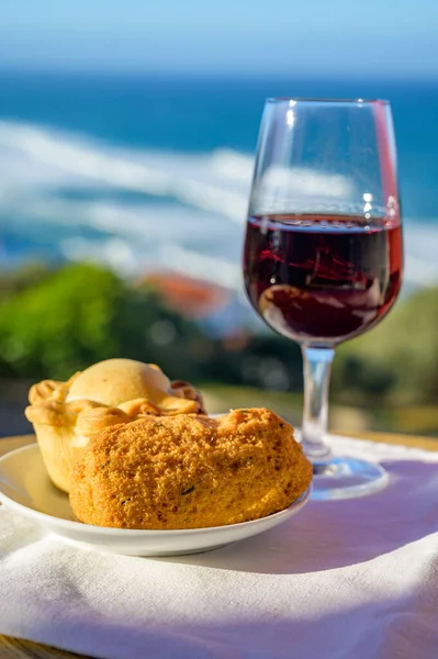 Portugeese food and drink, oven baked chicken pie empada de frango and codfish croquette pastais de bacalhau, glass of porto wine, served outdoor with view on blue Atlantic ocean near Sintra in Lisbon area, Portugal, close up