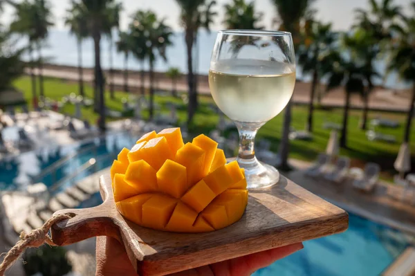 Stock image Fresh ripe sweet yellow mango tropical fruit served outdoor with glass of cold white wine and view on palm trees and blue pool and sea