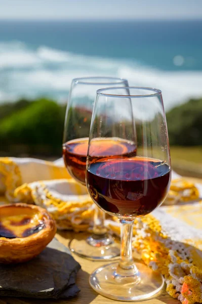 Portugal\'s traditional food and drink, glass of porto wine and muscatel de setubal, sweet dessert Pastel de nata egg custard tart pastry served with view on blue Atlantic ocean near Sintra in Lisbon area, Portugal