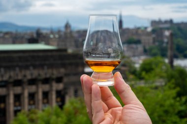 Hand holding glass of single malt scotch whisky and view from Calton hill to park and old parts of Edinburgh city in rainy summer day, Scotland, UK clipart