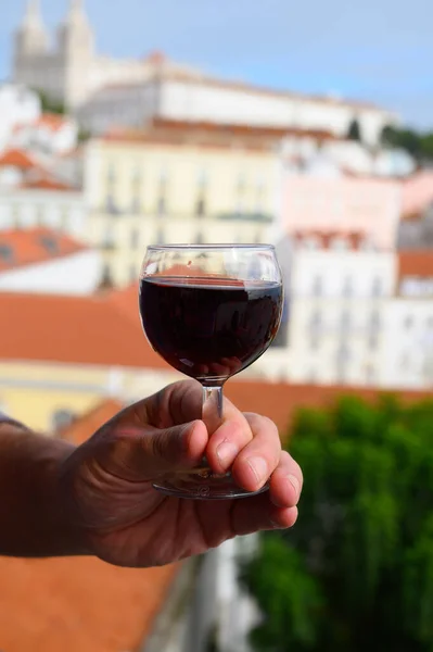 Hand with glass of ruby porto Portuguese wine in outdoor cafe at view point on colorful old part of Lisbon city, Portugal