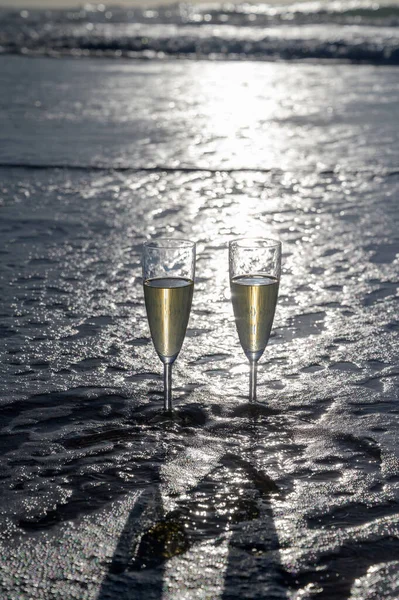 Glasses of cava or champagne sparkling wine on white sandy ocean beach with water waves on sunset in sunlights with shadows