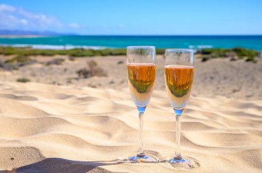 Two glasses of rose champagne or cava sparkling wine served on the white sandy tropical beach with dunes and blue ocean, romantic vacation, winter sun on Fuerteventura, Canary, Spain