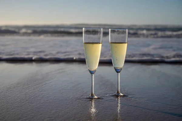 Glasses of cava or champagne sparkling wine on white sandy ocean beach with water waves on sunset in sunlights with shadows
