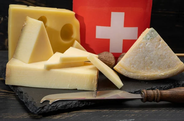 Assortment of Swiss cheeses Emmental or Emmentaler medium-hard cheese with round holes, Gruyere, appenzeller used for traditional cheese fondue and gratin and flag of Switzerland on dark background