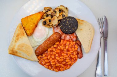 White board with full English breakfast with bacon, fried egg, beans, tomato, roasted sausage, black pudding, scons, hash browns and fried mushrooms close up