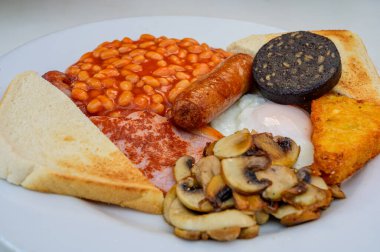 White board with full English breakfast with bacon, fried egg, beans, tomato, roasted sausage, black pudding, scons, hash browns and fried mushrooms close up