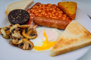 White board with full English breakfast with bacon, fried egg, beans, tomato, roasted sausage, black pudding, scons, hash browns and fried mushrooms close up