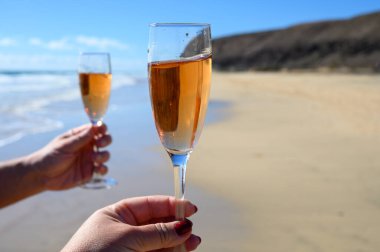 Beach party, hands with glass of rose champagne or cava sparkling wine with view on white sandy tropical beach and blue ocean water, romantic vacation, winter sun on Fuerteventura, Canary, Spain