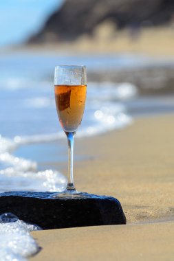 One glass of rose champagne or cava sparkling wine served on stone on white sandy tropical beach and blue ocean water, romantic vacation, winter sun on Fuerteventura, Canary, Spain