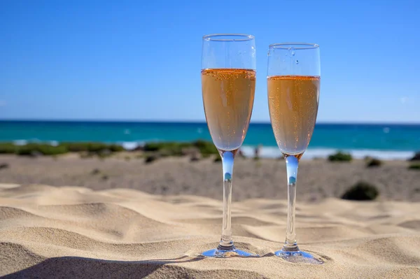 stock image Two glasses of rose champagne or cava sparkling wine served on the white sandy tropical beach with dunes and blue ocean, romantic vacation, winter sun on Fuerteventura, Canary, Spain