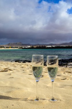 Two glasses of white champagne or cava sparkling wine served on white sandy tropical beach and blue ocean, romantic vacation, winter sun on Fuerteventura, Il Cotillo, Canary, Spain