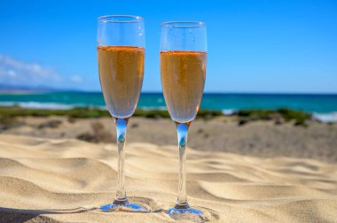Two glasses of rose champagne or cava sparkling wine served on the white sandy tropical beach with dunes and blue ocean, romantic vacation, winter sun on Fuerteventura, Canary, Spain