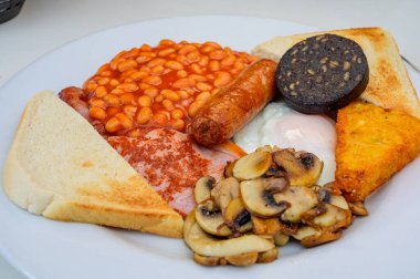 White board with full English breakfast with bacon, fried egg, beans, tomato, roasted sausage, black pudding, scons, hash browns and fried mushrooms close up