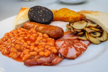 White board with full English breakfast with bacon, fried egg, beans, tomato, roasted sausage, black pudding, scons, hash browns and fried mushrooms close up