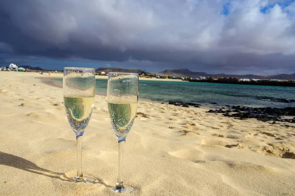 stock image Two glasses of white champagne or cava sparkling wine served on white sandy tropical beach and blue ocean, romantic vacation, winter sun on Fuerteventura, Il Cotillo, Canary, Spain