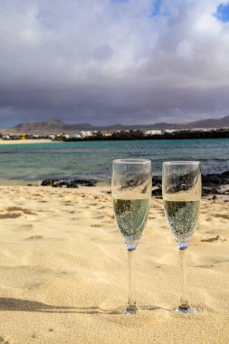 Two glasses of white champagne or cava sparkling wine served on white sandy tropical beach and blue ocean, romantic vacation, winter sun on Fuerteventura, Il Cotillo, Canary, Spain