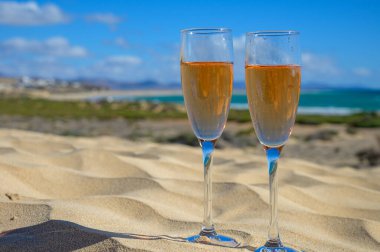 Two glasses of rose champagne or cava sparkling wine served on the white sandy tropical beach with dunes and blue ocean, romantic vacation, winter sun on Fuerteventura, Canary, Spain