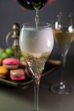 Pouring of sparkling white wine champagne or cava with bubbles in glasses and sweet dessert colorful macarons biscuits on background