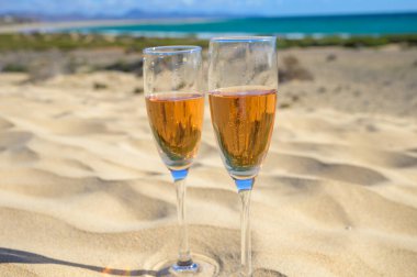 Two glasses of rose champagne or cava sparkling wine served on the white sandy tropical beach with dunes and blue ocean, romantic vacation, winter sun on Fuerteventura, Canary, Spain