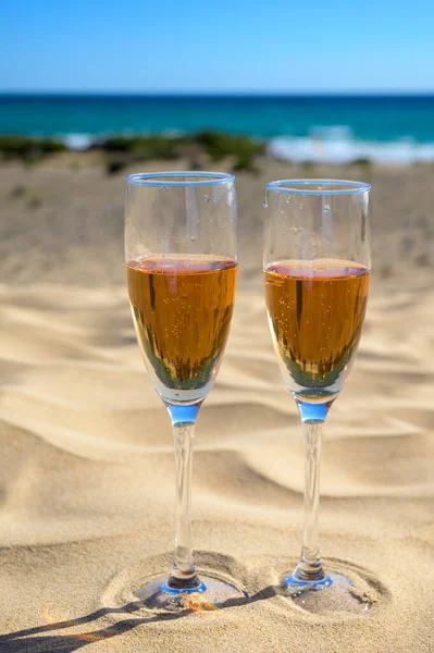 Two glasses of rose champagne or cava sparkling wine served on the white sandy tropical beach with dunes and blue ocean, romantic vacation, winter sun on Fuerteventura, Canary, Spain