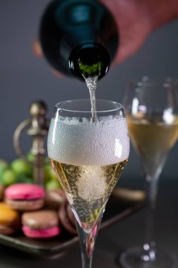 Pouring of sparkling white wine champagne or cava with bubbles in glasses and sweet dessert colorful macarons biscuits on background