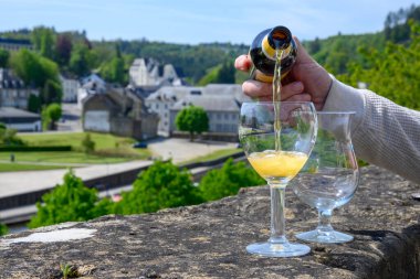 Pouring of blond strong Belgian abbey beer in to glass in sunny summer day with nice view on old town Bouillon, Belgium clipart