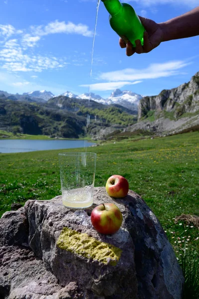 stock image Pouring of natural Asturian cider made from fermented apples from green bottle and high height with view on Covadonga lake and tops of Picos de Europa mountains, North of Spain