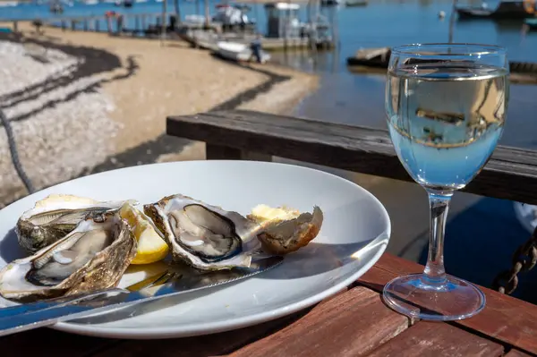 stock image Eating of fresh live oysters with glass of white wine at farm cafe in oyster-farming village, with view on boats and water of Arcachon bay, Cap Ferret peninsula, Bordeaux, France in sunny day