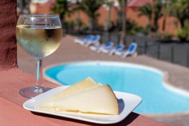 İspanyol sert manchegosu, inek, koyun ve keçi peyniri, arka planda bir bardak soğuk beyaz şarap ve mavi yüzme havuzu