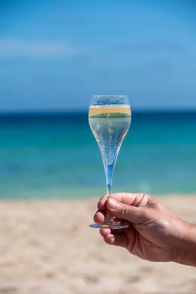 Hand with glass of cava or champagne sparkling wine on vacation, Dunes Corralejo Grand sandy beach, Fuerteventura, Canary islands, blue ocean