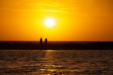 Erkek ve kadın kalıp okyanus sahilinde gün batımını izliyorlar, turuncu gökyüzü, tatildeki insanların siluetleri, Kanarya adaları.