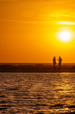 Erkek ve kadın kalıp okyanus sahilinde gün batımını izliyorlar, turuncu gökyüzü, tatildeki insanların siluetleri, Kanarya adaları.