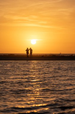 Erkek ve kadın kalıp okyanus sahilinde gün batımını izliyorlar, turuncu gökyüzü, tatildeki insanların siluetleri, Kanarya adaları.