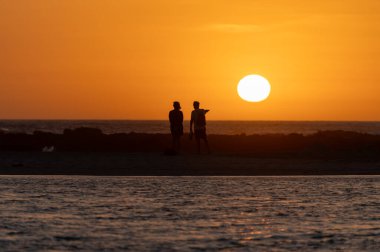 Erkek ve kadın kalıp okyanus sahilinde gün batımını izliyorlar, turuncu gökyüzü, tatildeki insanların siluetleri, Kanarya adaları.