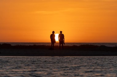 Erkek ve kadın kalıp okyanus sahilinde gün batımını izliyorlar, turuncu gökyüzü, tatildeki insanların siluetleri, Kanarya adaları.