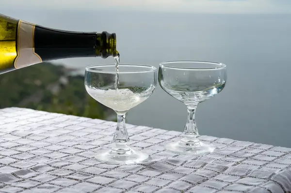 stock image Pouring of cold sparkling wine, cava or champagne in two coupe glasses on table with sea view