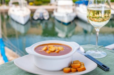 Homemade tasty fish or bisque soup with croutons served outdoor with glass of cold white wine and view on yacht boats and houses of Port Grimaud, French Riviera, France. Food of Provence clipart