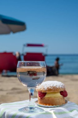 Saint-Tropez 'deki kumlu plajda krema ve taze kırmızı böğürtlenlerle dolu tarte tropezienne kekiyle öğle yemeği. Bir bardak gül şarabı, güneşli Fransız Rivierasında yaz tatili, Fransa.