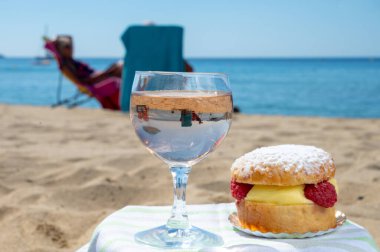 Lunch on sandy beach in Saint-Tropez with cake Tarte tropezienne filled brioche with cream and fresh red berries and glass of rose wine, summer vacation on sunny  French riviera, France clipart