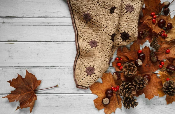 Warm, cozy handmade blanket, with autumn leaves, cones and chestnuts. Beautiful fall flat lay composition on white wooden table, with copyspace.