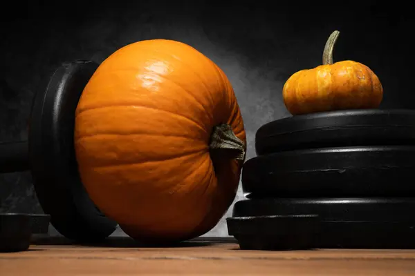 Dumbbell barbell with orange pumpkin as a weight plate. Gym weightlifting workout and sport training concept. Healthy fitness lifestyle autumn or fall composition for Halloween or Thanksgiving.