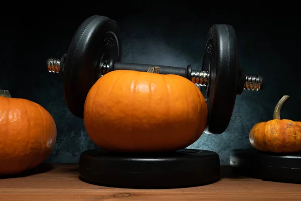 Orange autumn pumpkin lifting heavy barbell dumbbell. Weightlifting fall idea for Halloween or Thanksgiving. Gym weight workout and sport training concept. Healthy lifestyle composition.